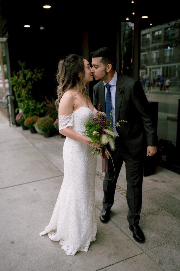 A newlywed couple kissing on a city sidewalk, holding a bouquet of flowers.